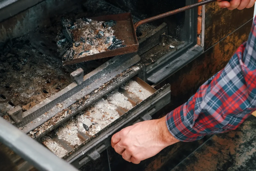 A person in a plaid shirt is cleaning ashes from a wood stove or fireplace chimney, using a metal shovel to scoop ashes from the ashtray compartment.