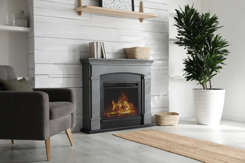 A modern living room with a gray armchair, a black fireplace with a glowing flame reminiscent of a Franklin stove, a large potted plant, and neutral decor on a light wood floor with white paneled walls.
