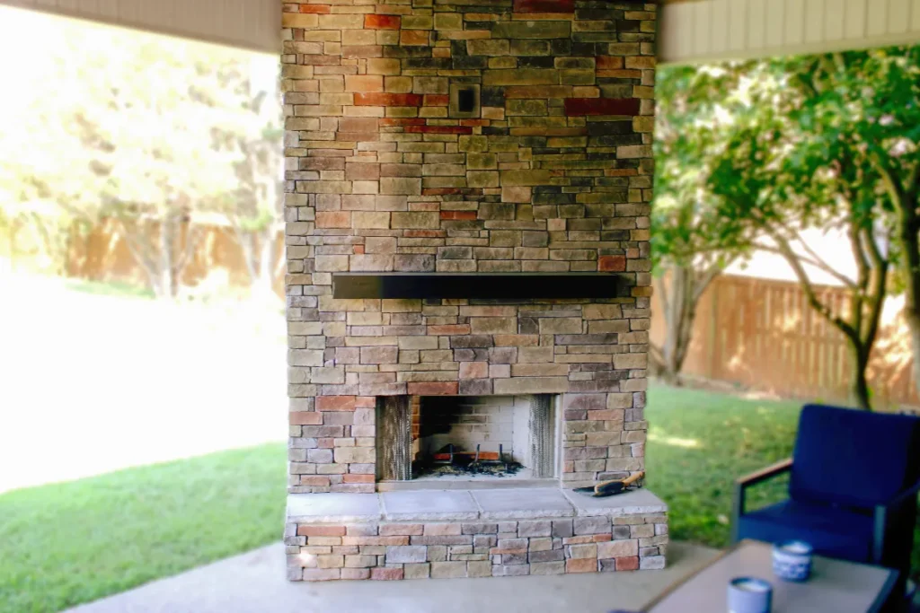 Stone outdoor fireplace with a black mantel and chimney under a covered patio in Nashville, surrounded by patio furniture and green trees in the background.